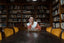 A woman at a long dark table with gold velvet chairs looks down at the table, with filled bookshelves behind her