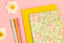 A floral notebook sits atop a yellow notebook, next to colorful pens and daisies on a pink table