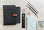 A planner in a wool sleeve, on a wooden desk next to office supplies like pencils, tape, a pen, envelopes, and paper clips.