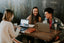 3 people with laptops and drinks sit around a wood table smiling and laughing