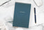 A dark blue gemstone spiral one-on-one pad on a white desk next to a pen
