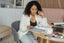 A woman sits at a desk reading a magazine while talking on the phone