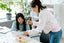 Two women smile and work on a white desk looking at a sheet of paper together
