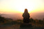 A woman sits outside in front of a sunset over hills