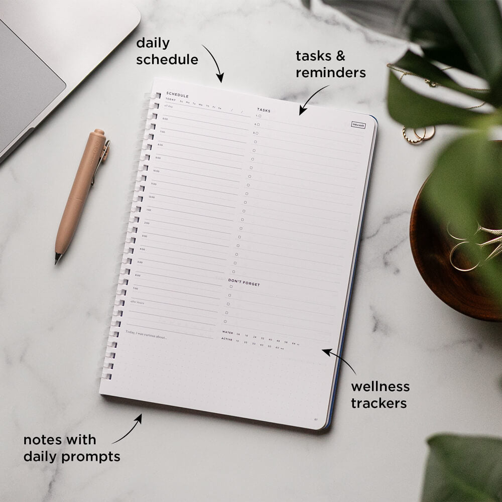 Planner with sections labeled for daily schedule, tasks & reminders, wellness trackers, and notes with daily prompts on a marble surface.